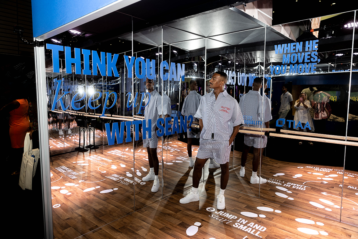 Sean Bankhead is wearing a matching blue and white striped shirt with all white shoes and stands in a mirrored dance studio with wooden floors. There is blue vinyl writing on the walls that says, “Think you can keep up with Sean?” and “When he moves, you move.” “#RetireInequality” and the TIAA logo. 